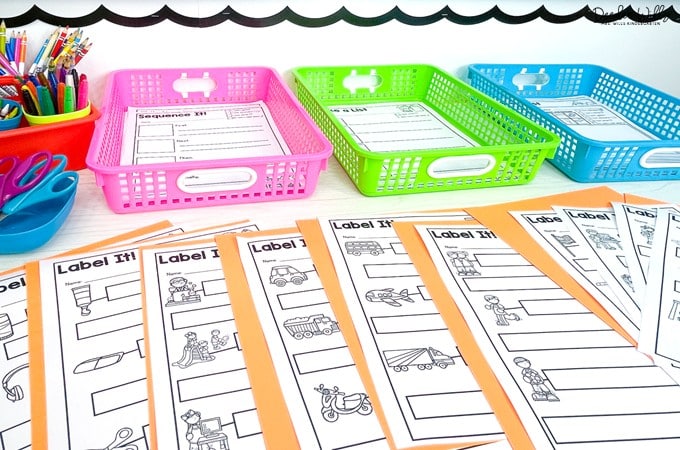 Kindergarten writing worksheets labeled “Label It!” spread on a table with colorful baskets holding completed pages and supplies.