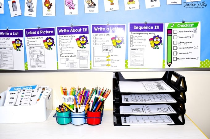 Organized kindergarten writing center table with writing posters, sight word cards, cups of pencils, and trays of worksheets.