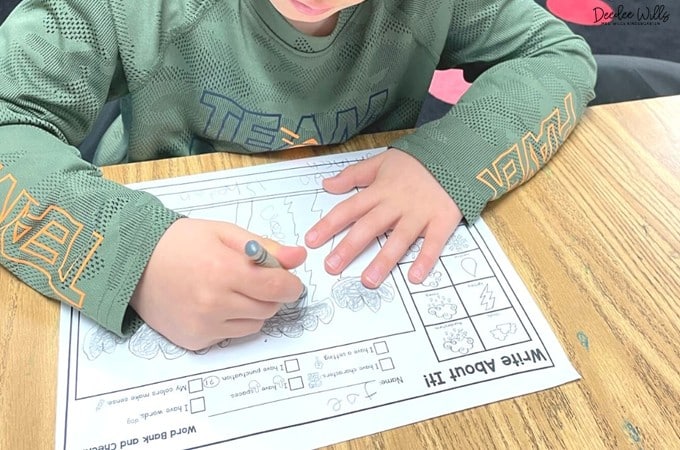 Student uses a crayon to complete a kindergarten writing worksheet at a classroom table, focusing on drawing and labeling.