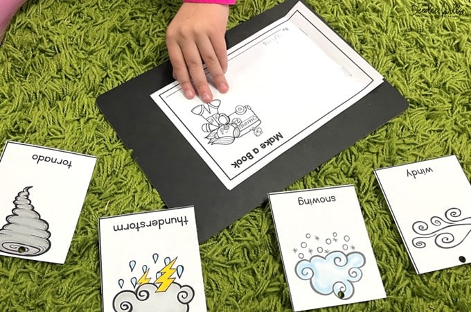 Child works on a weather writing page on a green carpet, surrounded by illustrated cards labeled tornado, thunderstorm, snowing, and windy.