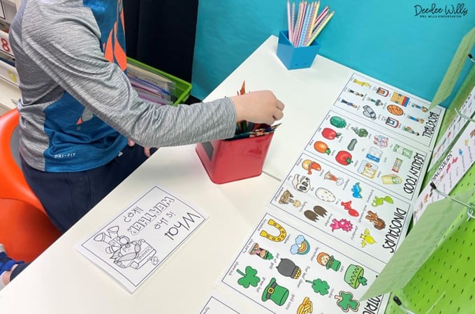 Student works at a kindergarten writing center, choosing supplies while using picture word charts and themed writing materials on the table.